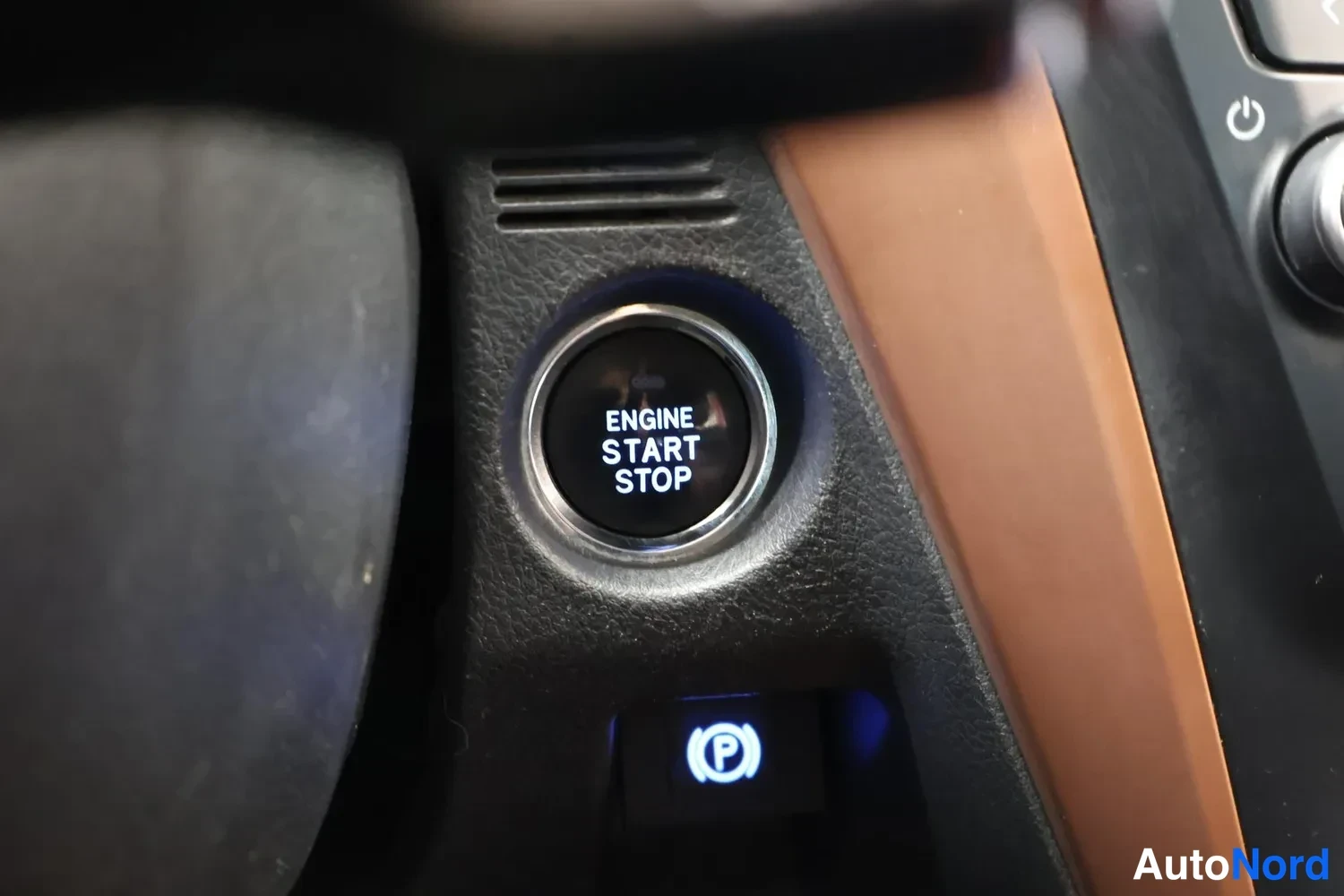 Yksityiskohta: Sisätilakuva: moottorin käynnistys-/sammutuspainike (ENGINE START STOP) ja sen alla sinisellä valaistu electronic parking brake -painike.