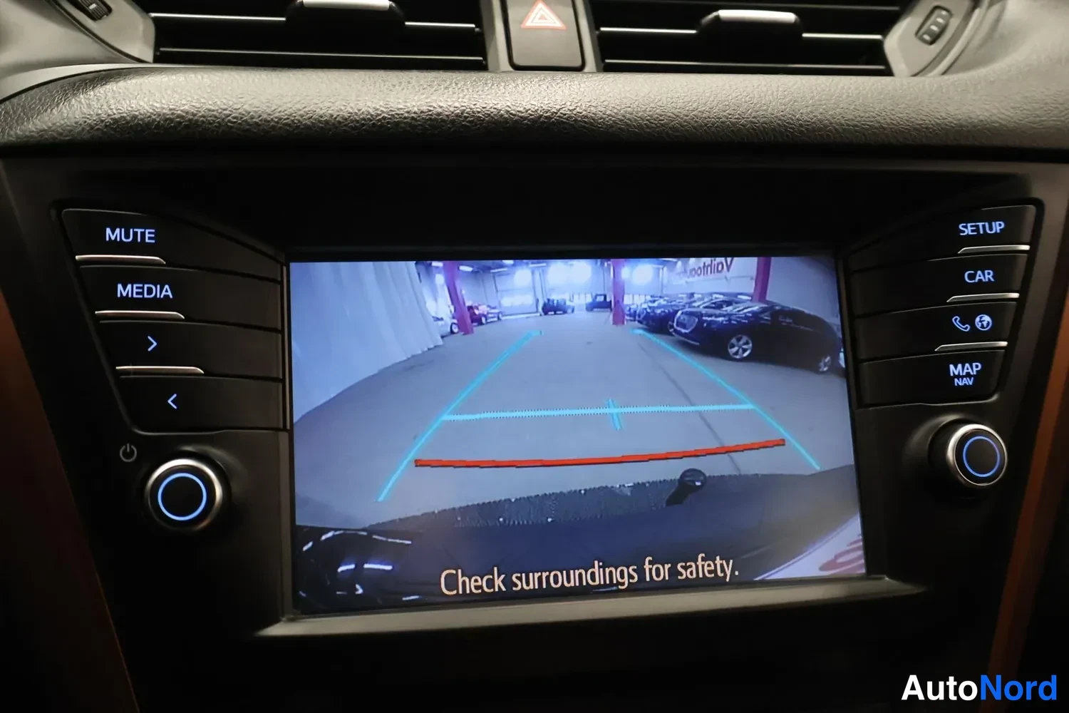 Yksityiskohta: Viihdejärjestelmän näyttö näyttää peruutuskameran kuvan parkitsema-autoluotoilla ja tekstillä Check surroundings for safety.