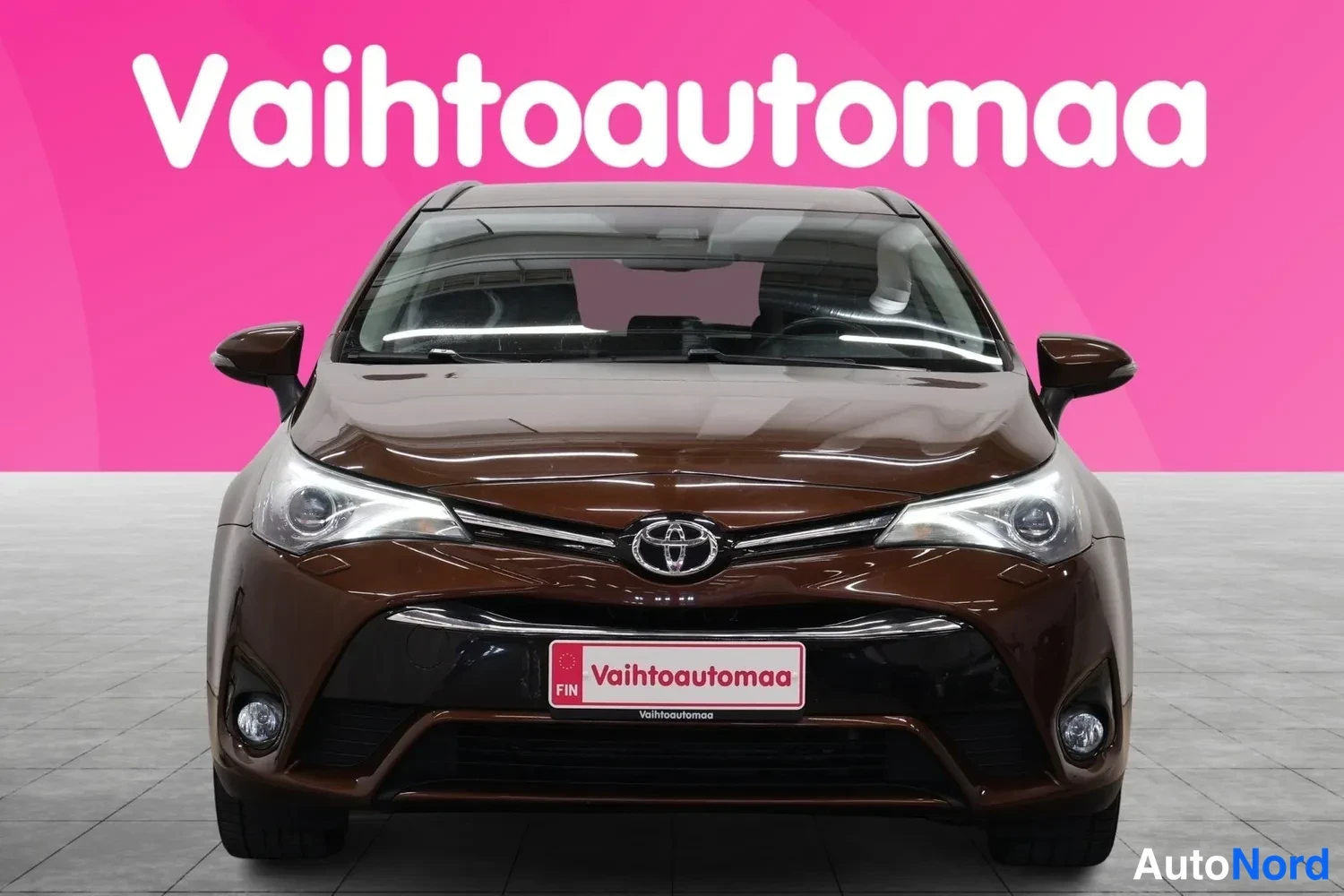 Edestä: Ruskean farmariauton suora etunäkymä, Toyota-merkki ja etupuskuri näkyvissä, pinkki tausta.
