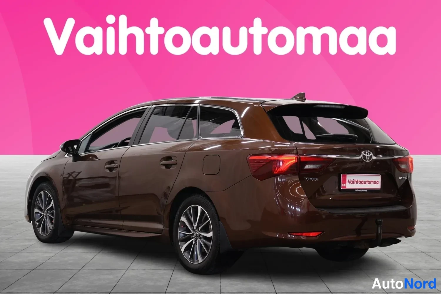 Takaviisto: Ruskean Toyota Avensis -farmarin takaosasivukulma vasemmalta, takavalojen ja Puolan rekisterikilven kanssa pinkillä taustalla.