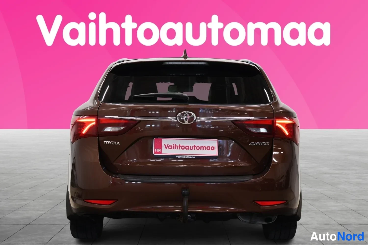 Takaa: Ruskean Toyota Avensis -farmarin takanäkymä: Toyota-merkki, avensis-teksti ja Vaihtoautomaa-kilpi pinkillä taustalla.