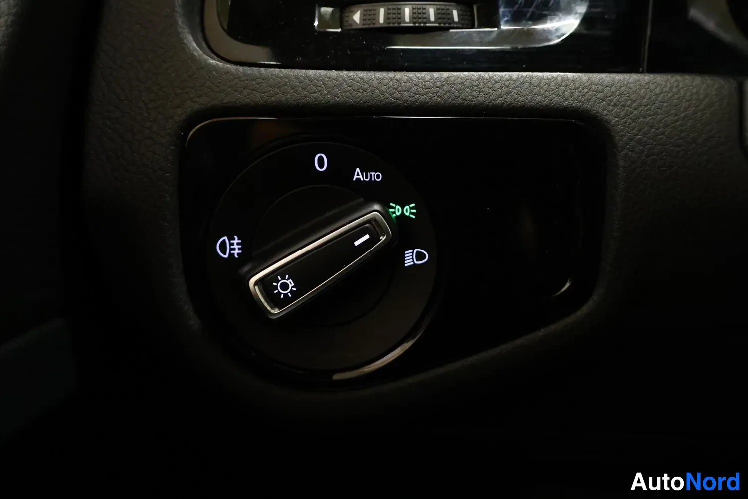 Yksityiskohta: Auton valokytkimen nuppi lähikuvassa asennossa, jossa on Auto-tila ja vinkkimerkintä; merkit 0, Auto ja sumuvalot erottuvat.