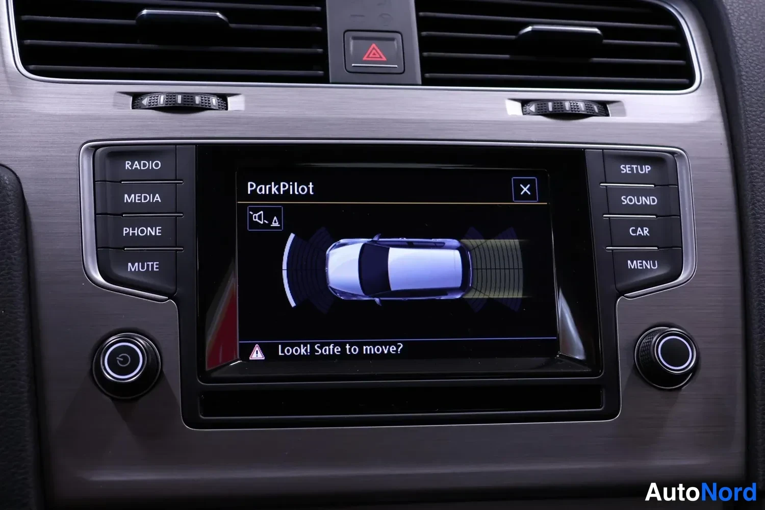 Muu: Volkswagenin kojelaudan radiopaneeli, jonka näytöllä on ParkPilot-tutkatnäkymä tekstillä Look! Safe to move?
