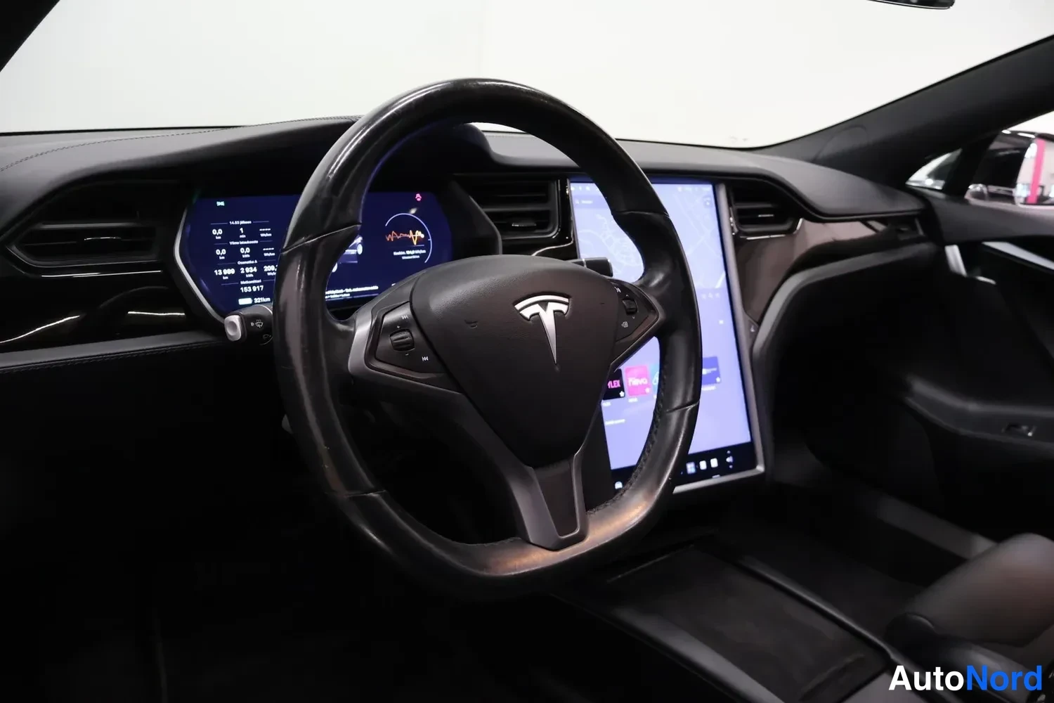 Sivulta: Ohjaamon etukuva: Tesla-logolla varustettu ohjauspyörä, digitaalinen mittaristo ja keskikonsolin suuri kosketusnäyttö.