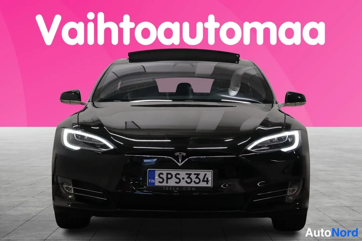 Edestä: Mustan Tesla Model S -sedanin suora etunäkymä, rekisterikilpi, taustana pinkki mainospinta.