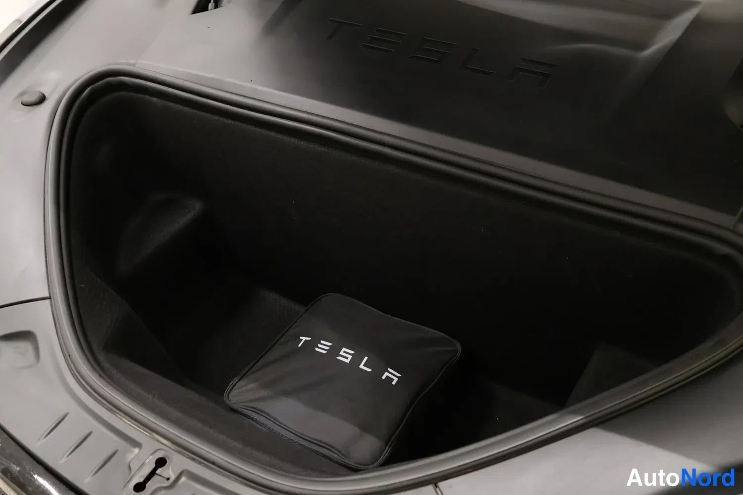Muu: Etutavaratila avattuna: tumma kokoomamatto ja musta vetoketjullinen säilytyspussi, jossa valkoinen TESLA-teksti; konepellin sisäpinnassa TESLA-kohokirjoitus.