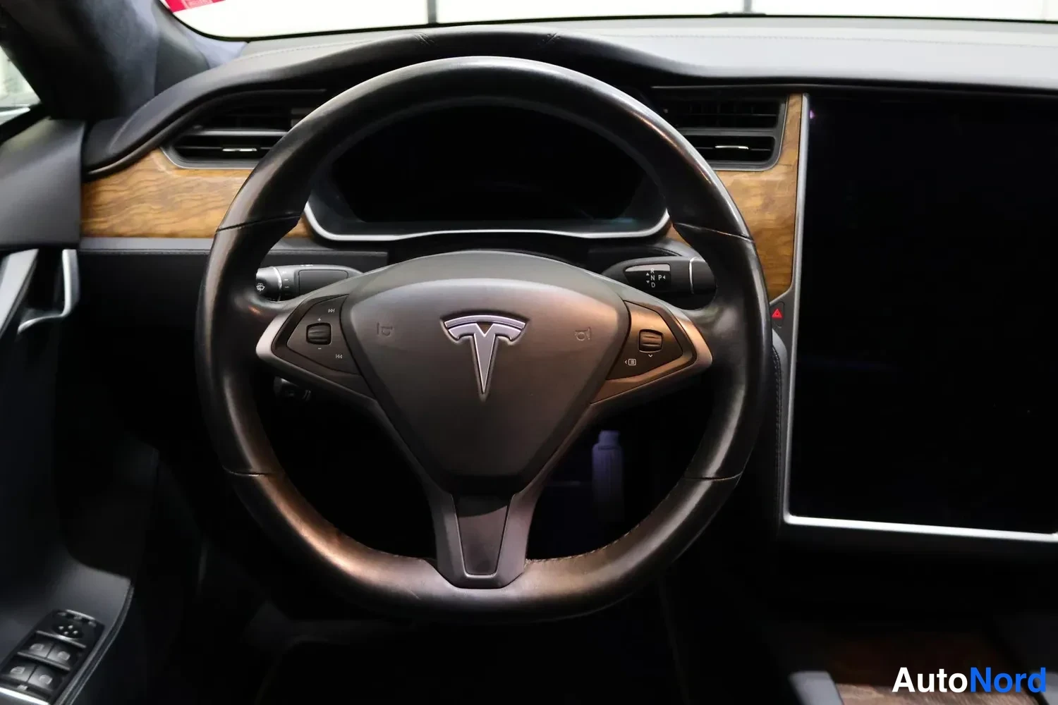 Muu: Ohjaamon näkymä: Tesla-merkkinen ohjauspyörä keskellä, puukuvioinen koriste paneeli ja suuri vaakatasoinen näyttö oikealla