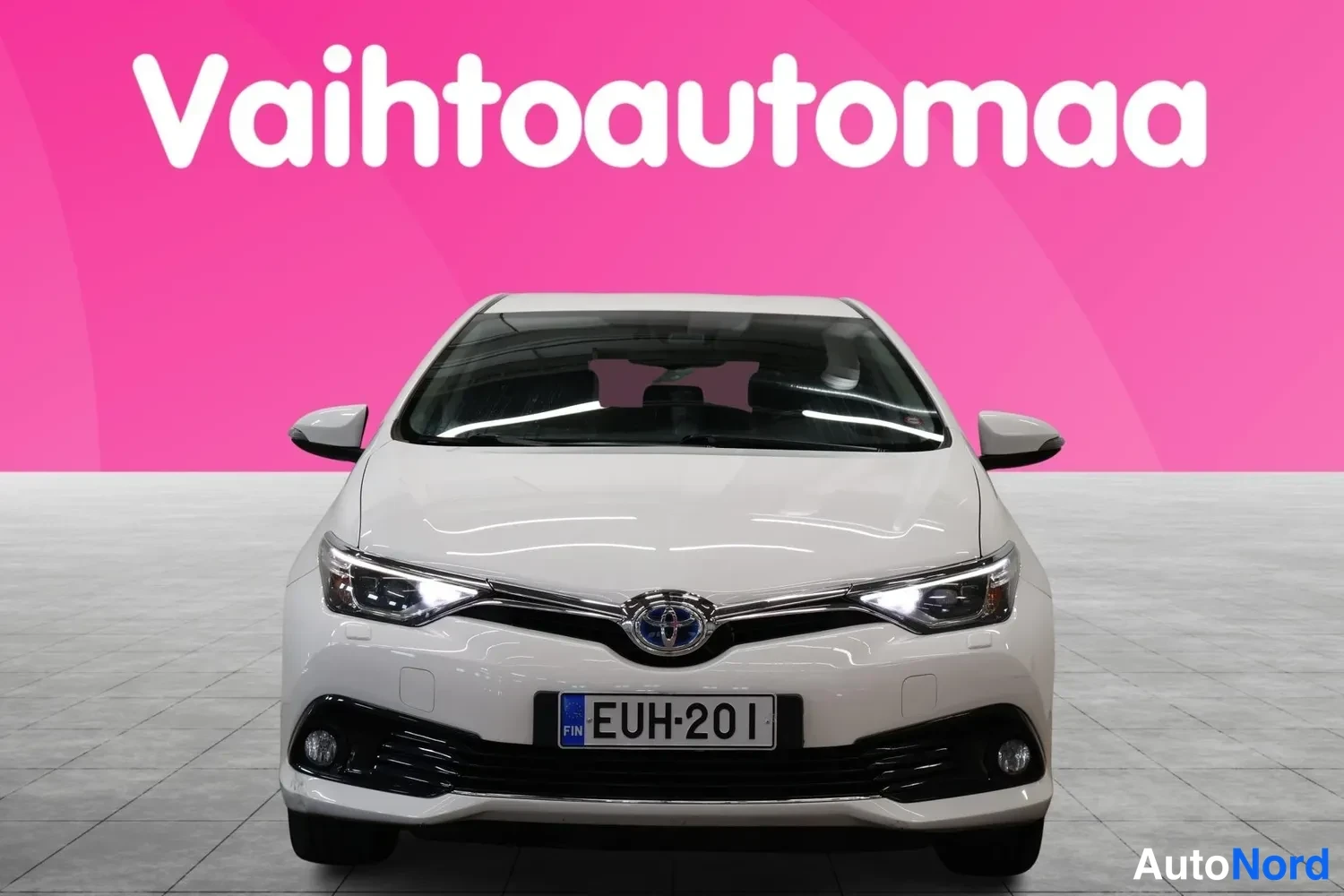 Edestä: Valkoinen henkilöauto suoraan edestäpäin kuvattuna, rekisterikilpi, pinkki tausta.