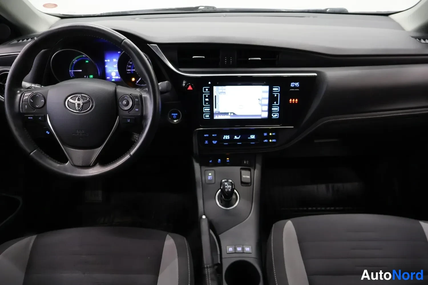 Yksityiskohta: Toyota-ohjauspyörä, hybridin mittaristo, kosketusnäyttö, automaattivaihteen valitsin ja tummat etuistuimet.
