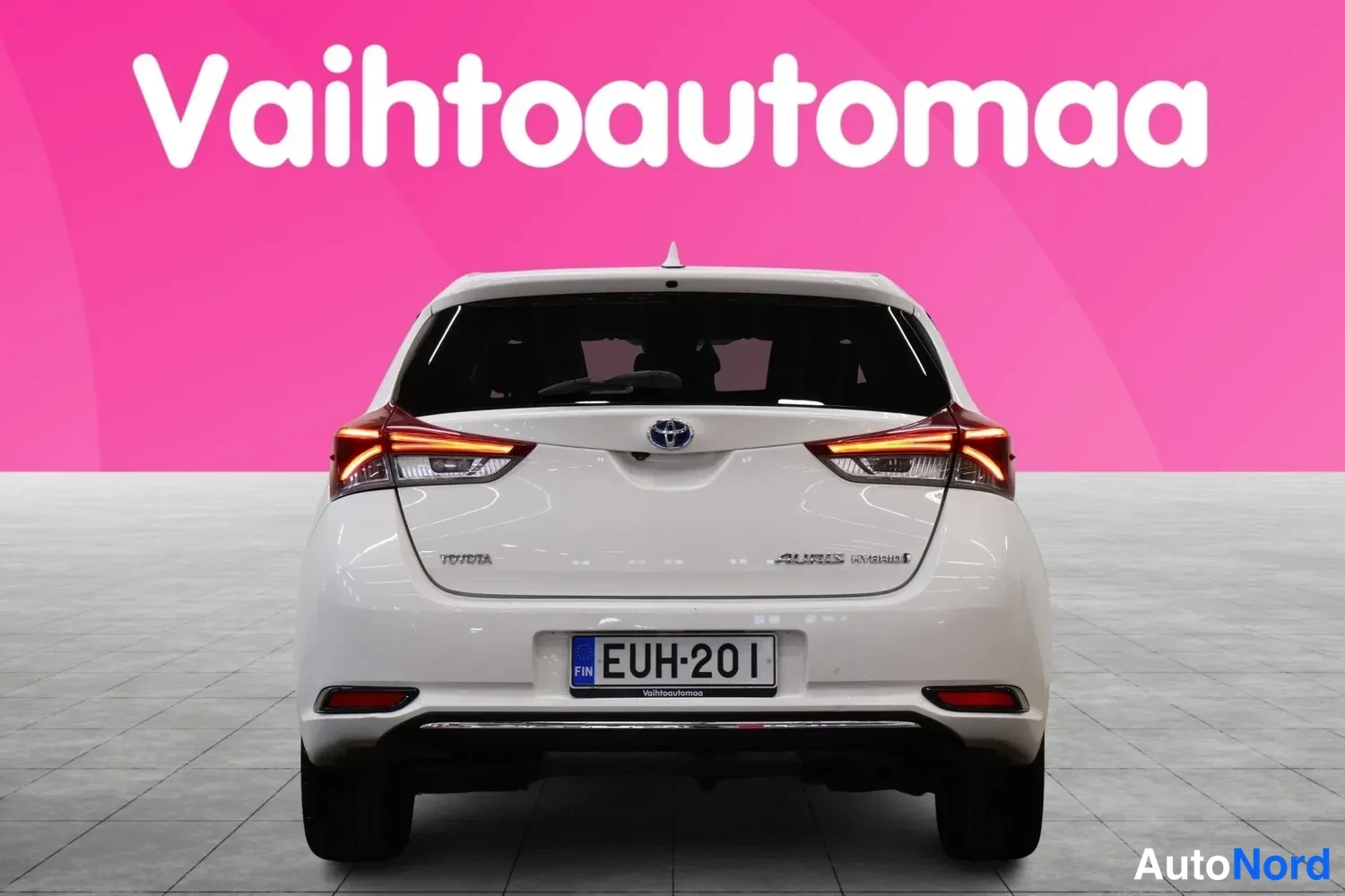 Takaa: Valkoisen hatchbackin takaosa suoraan takaa, punaiset takavalot, Toyota-merkki, Auris Hybrid-teksti ja -rekisterikilpi, vaaleansininen tausta.