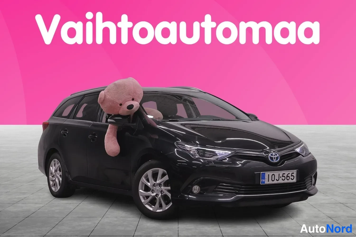 Etuviisto, nokka oikealle: Musta farmarivaihtoauto kolmivartalokuvaessa etunasta vasemmalla, rekisterikilpi 10J-565, taustana pinkki mainoslogo.