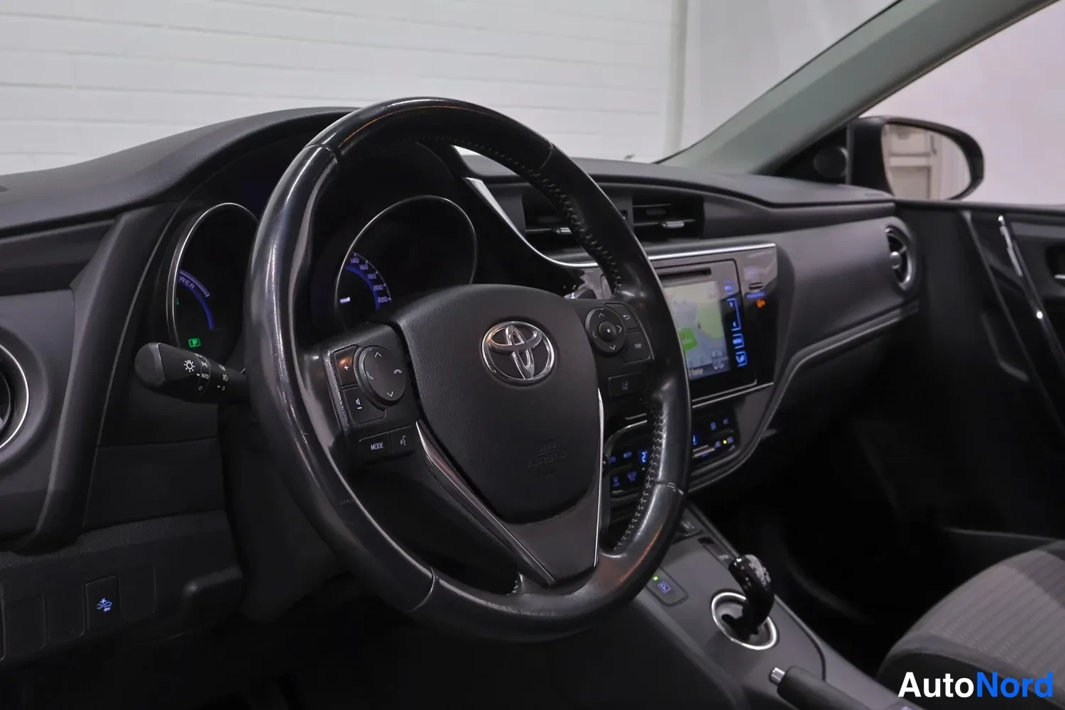 Sivulta: Ohjaamon näkymä: Toyota-merkkinen ohjauspyörä, kosketusnäyttö-navigaattori keskikonsolissa ja automaattivaihteen valitsin.