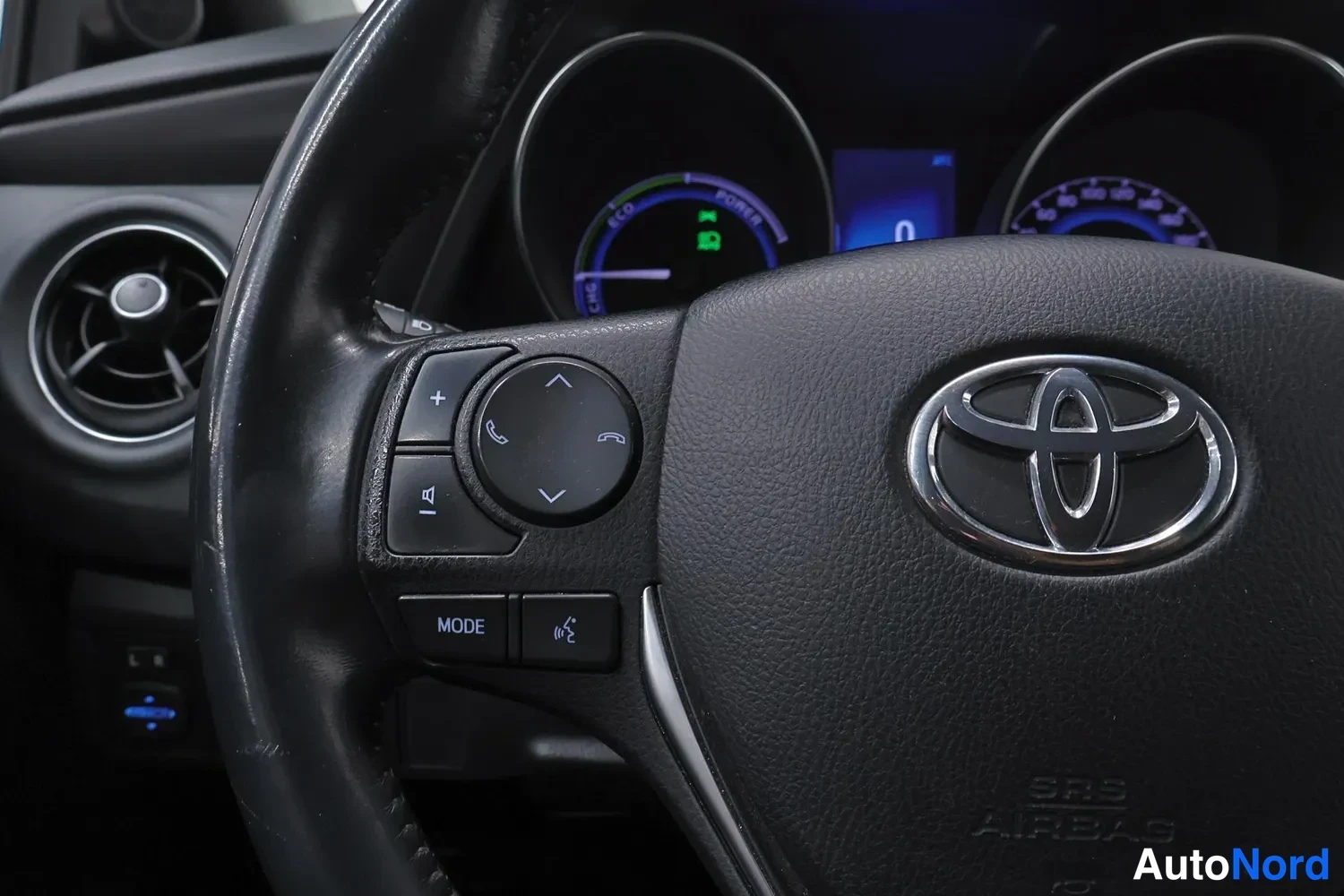 Etuviisto, nokka vasemmalle: Lähikuva Toyota-merkillä varustetusta ohjauspyörästä ja sen vasemmista painikkeista (MODE, puhelin- ja ääniohjausnappulat), taustalla hybridi- ja nopeu