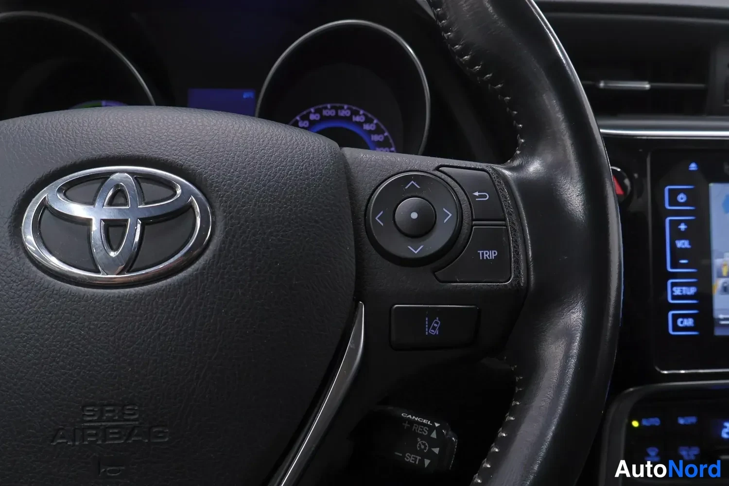 Etuviisto, nokka vasemmalle: Lähikuva ohjauspyörän oikean puolen painikkeista (TRIP, nuolinäppäimet, RES/SET-CANCEL-vipu), Toyota-emblem ja SRS AIRBAG -teksti näkyvissä, taustalla