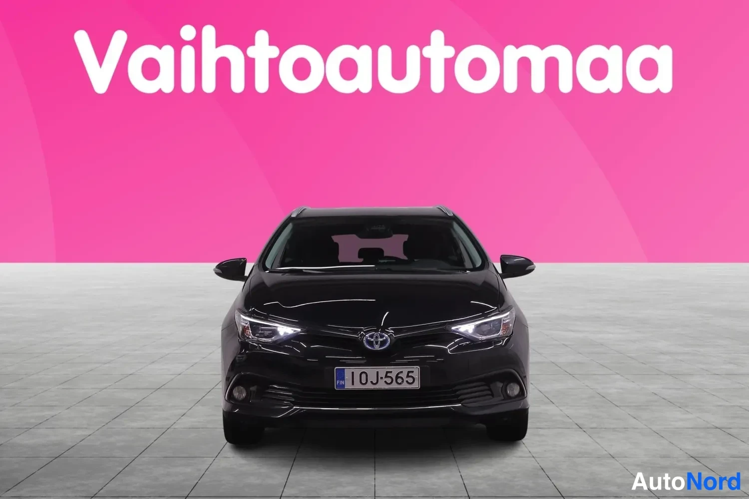 Edestä: Musta farmarivaihtoauto edestäpäin, rekisterikilpi 10J-565, taustana pinkki mainoslogo.