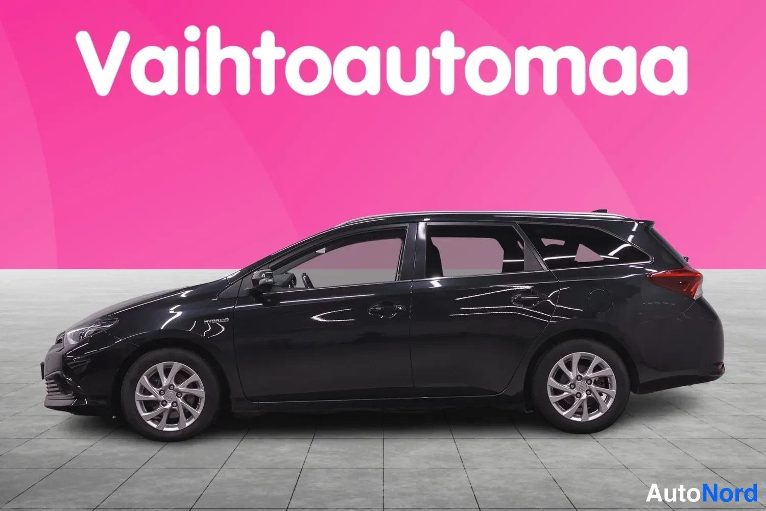 Etuviisto, nokka vasemmalle: Mustan Toyota Auris Hybrid -farmariauton sivuprofiili vasemmalta kuvattuna pinkin taustan edessä.