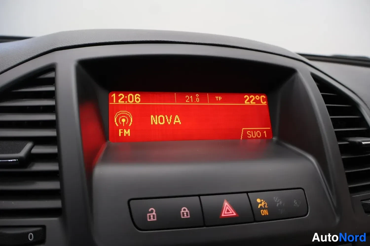 Yksityiskohta: Kojelaudan infonäyttö näyttää kellonajan 12:06, FM-radioaseman NOVA, lämpötilat 21,8 °C ja 22 °C sekä TP-merkinnän. Alla on lukituuspainikkeet, varoitusvilkkupa