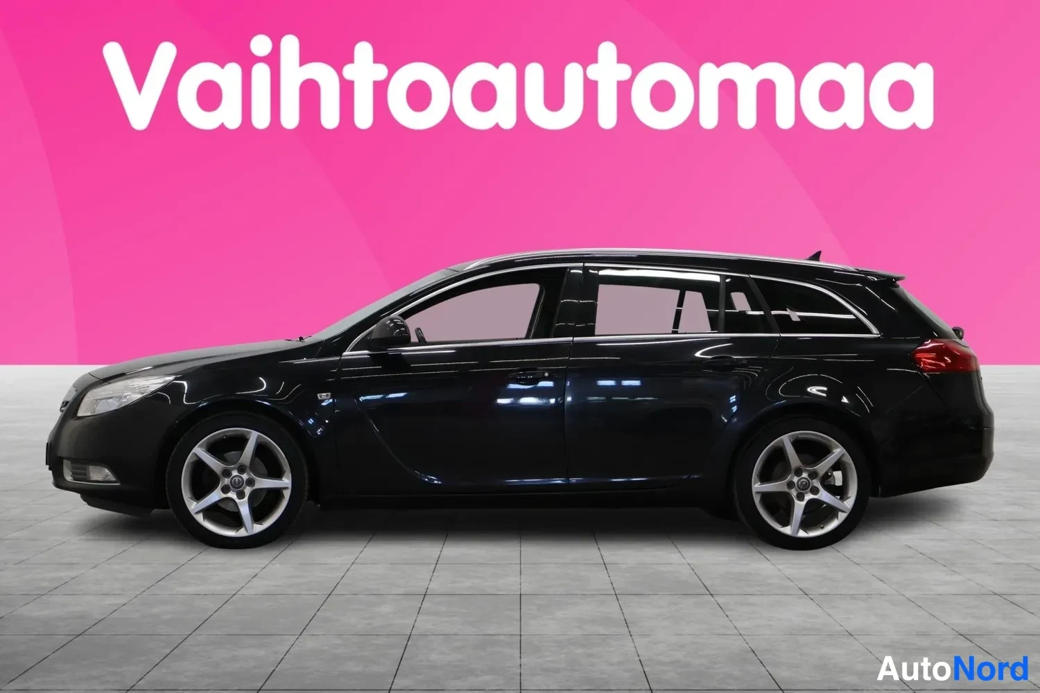 Etuviisto, nokka vasemmalle: Mustan farmariauto Opel Insignia Sports etuviistöstä vasemmalta, koko auto näkyvissä vaaleanpunaisella taustalla.