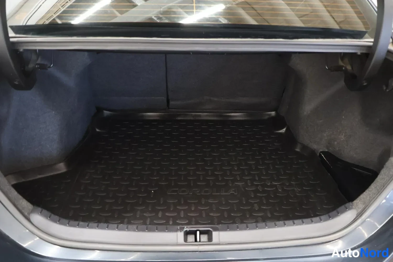 Muu: Auton avattu tavaratila: tumma pohjamatto ja teksti COROLLA maton pinnassa.