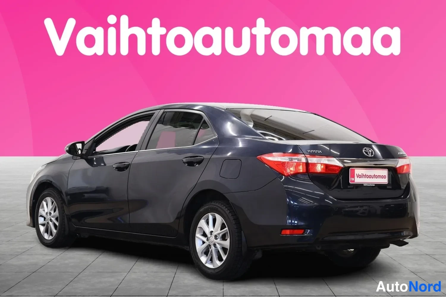 Takaviisto: Tummanharmaa Toyota Corolla -sedan takaviistosta oikealta; takavalot, Toyota-merkki ja Vahtoautomaa-kyltti näkyvät, etupyörä osittain.