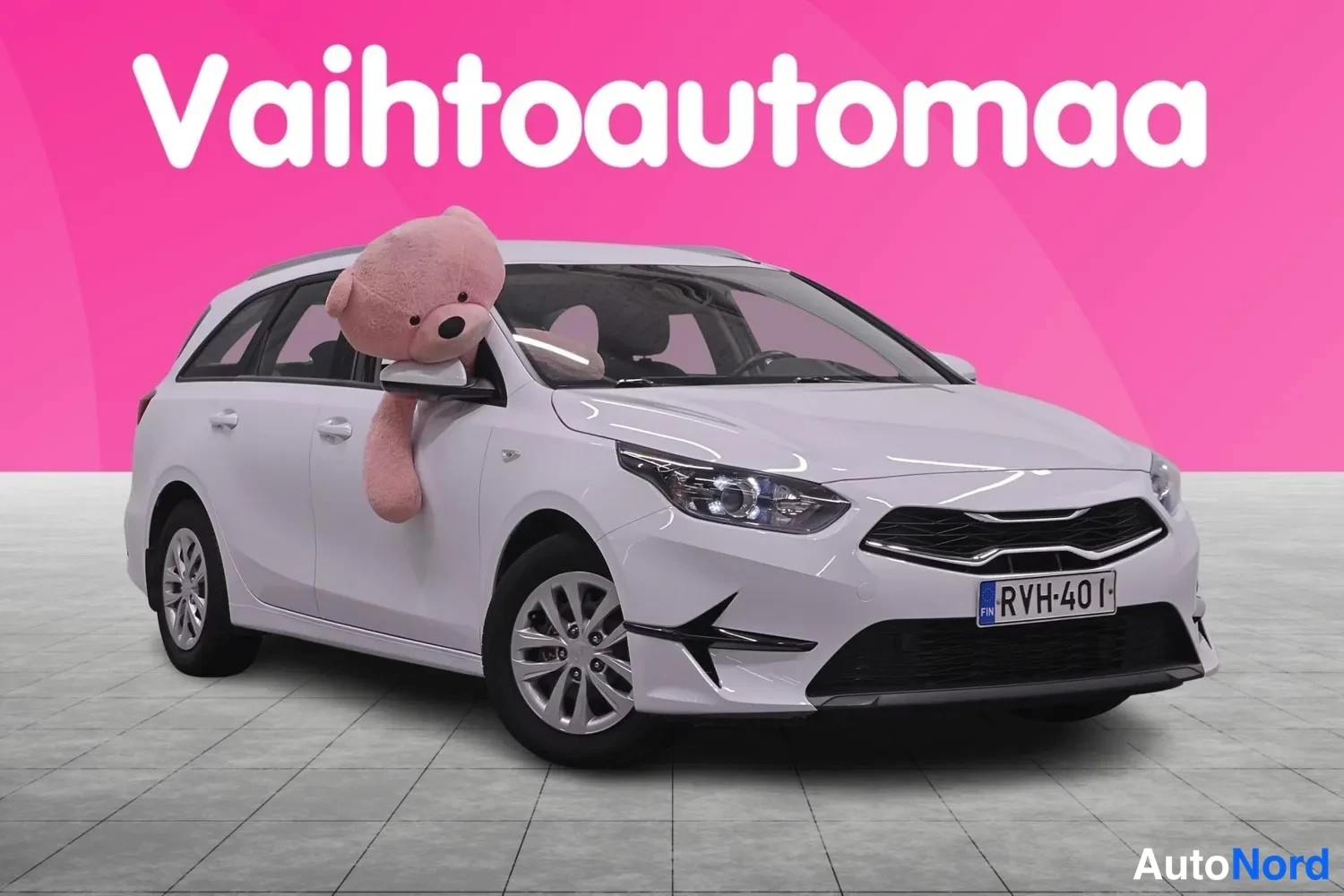 Etuviisto, nokka oikealle: Valkoinen farmariauto etuviistosta oikealta, rekisterikilpi, auton ovista kurkistaa vaaleanpunainen pehmolelu.