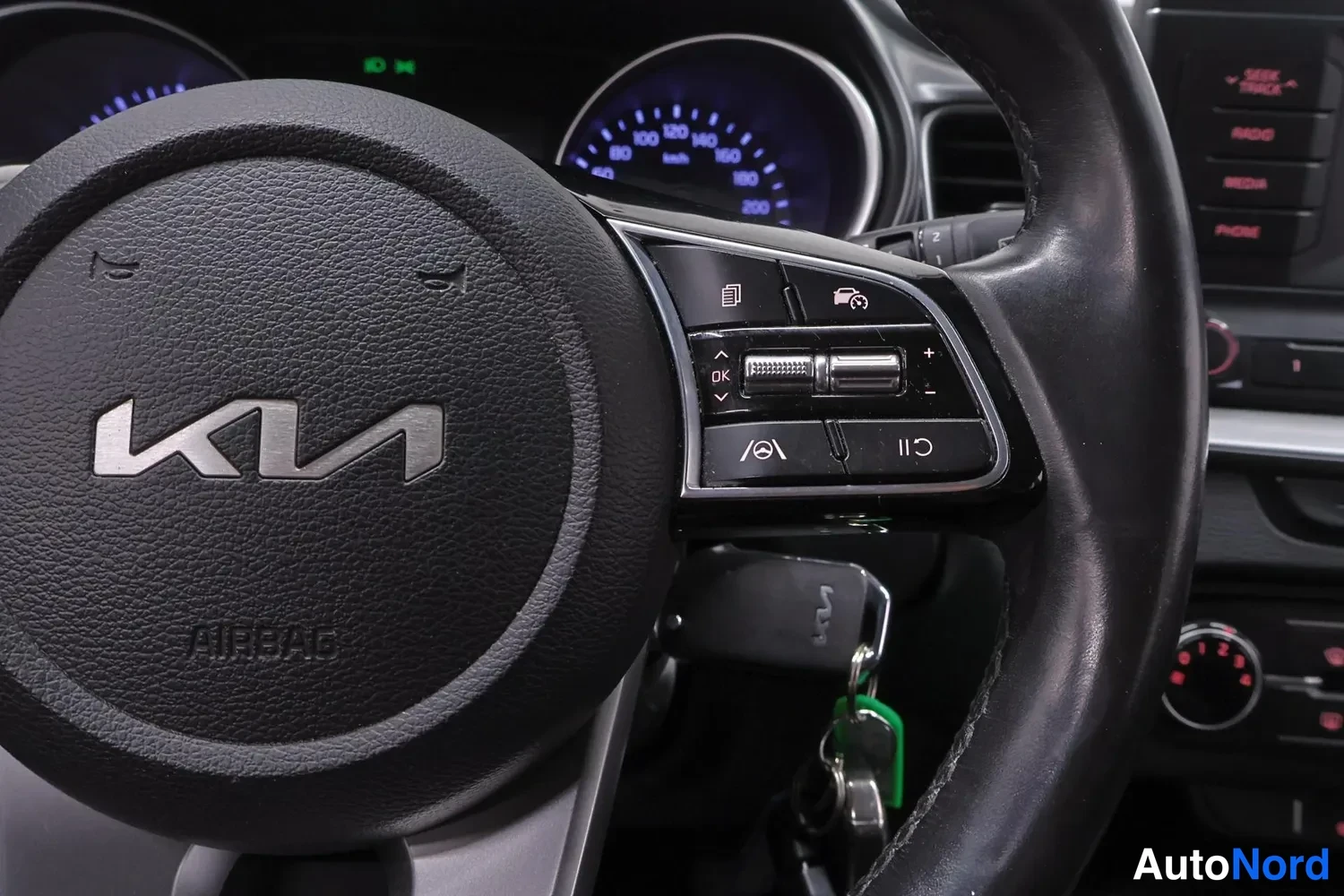 Sivulta: Ohjauspyörän lähikuva sisätiloissa: Kia-logo ja AIRBAG-teksti ohjauspyörässä, oikealla monitoiminapit vakionopeudensäädin- ja kaistavahti-ikoneilla, taustalla m