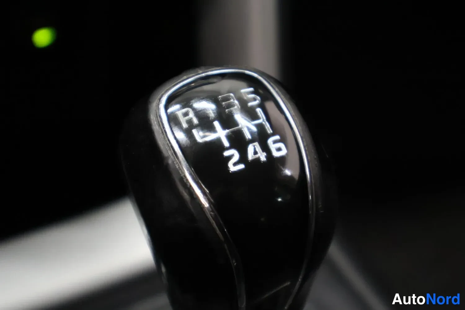 Yksityiskohta: Close-up of a manual gear shift with a black knob. The shift pattern is visible on the top face, indicating a 6-speed transmission (R 1-3-5 across the top row a