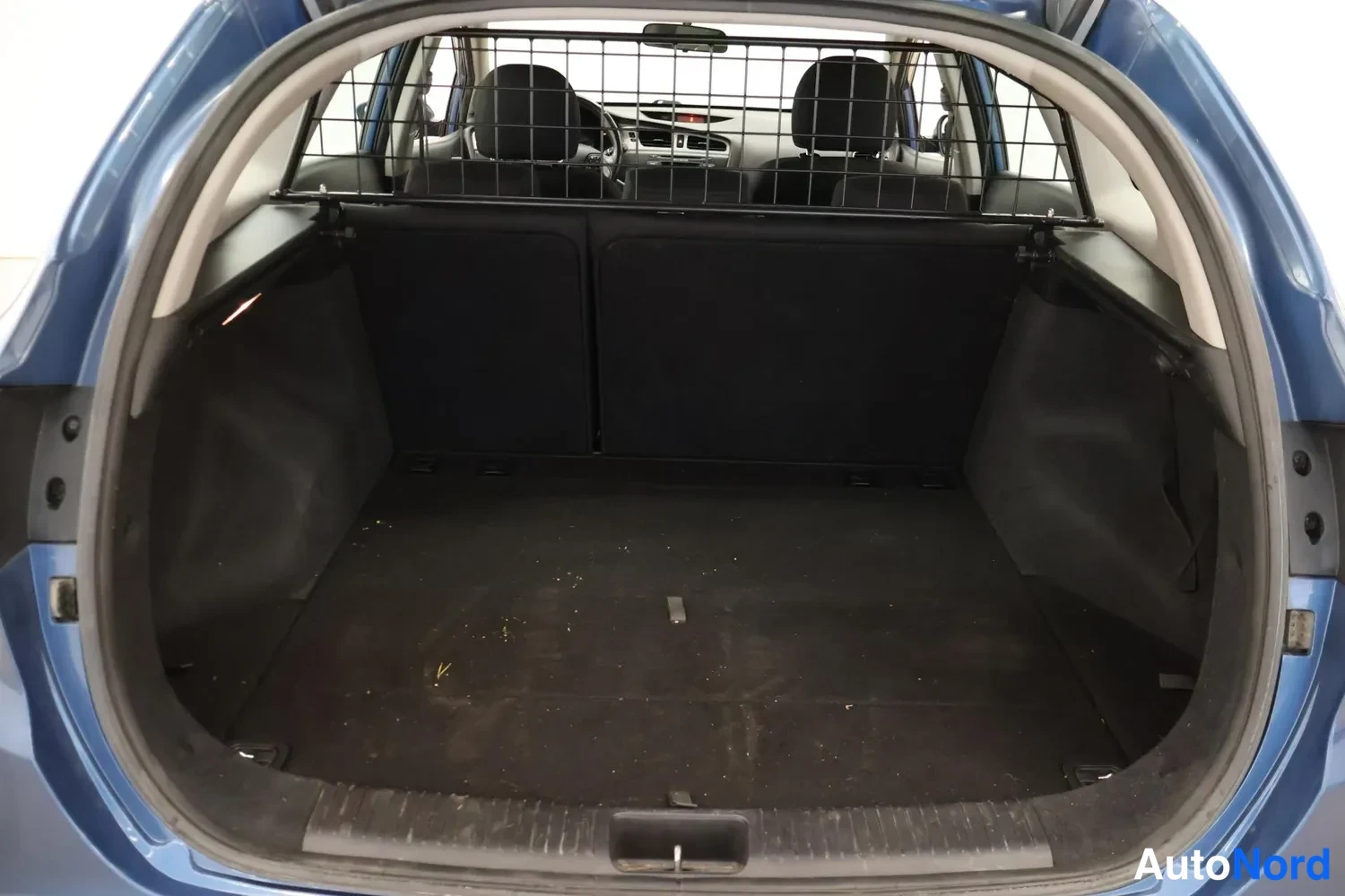 Tavaratila: Open trunk of a blue car. A metal cargo barrier separates the trunk space from the rear seats. Black luggage mats are installed in the boot floor.