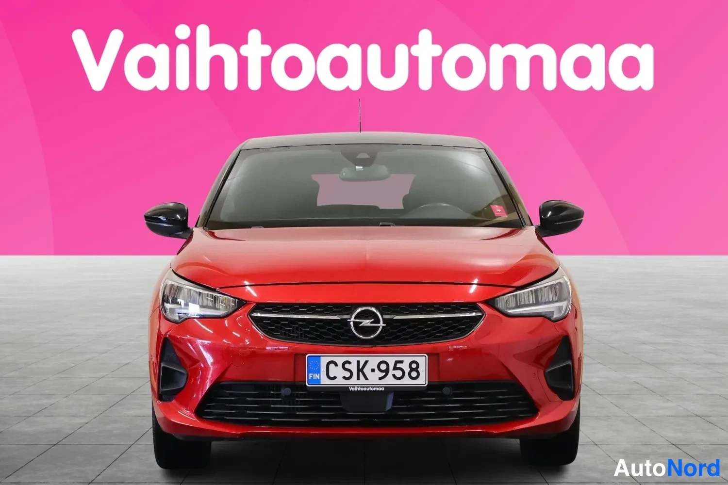 Edestä: Punaisen henkilöauton etunäkymä, Opel-merkki keulassa ja rekisterikilpi, pinkki tausta.