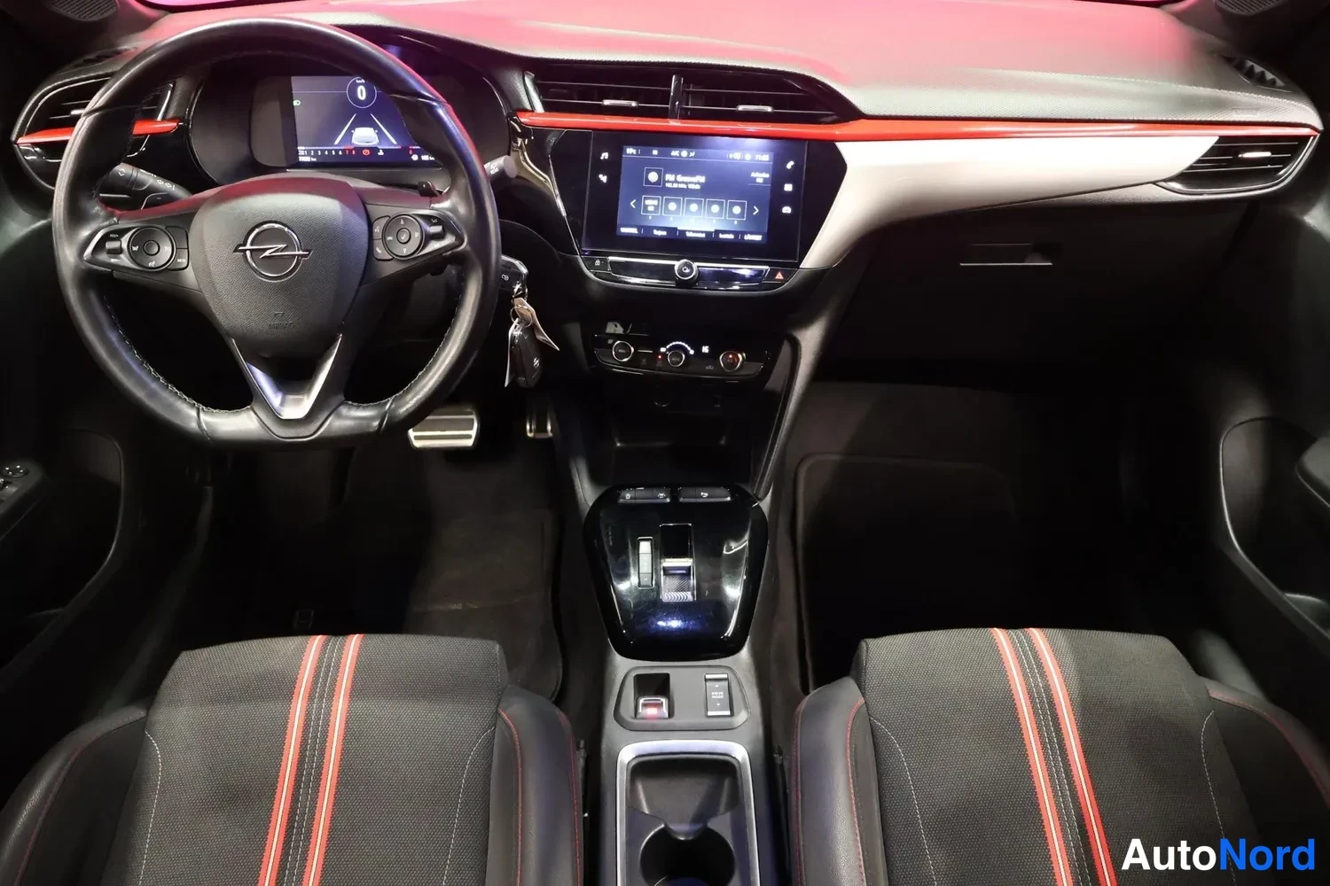 Etuviisto: Ohjaamo ylhäältä kuvattuna: Opel-ohjauspyörä, infotainment-näyttö ja automaattivaihteiston valitsin, istuimissa punaisia raidoita.