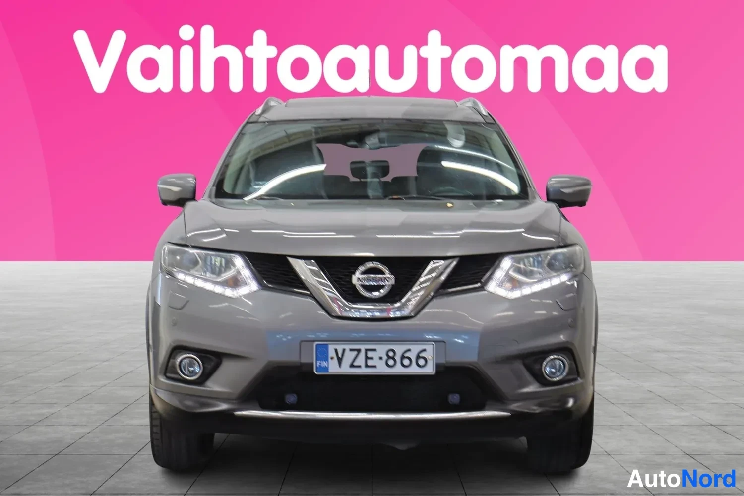 Edestä: Harmaan Nissan X-Trailin etupää suoraan edestä kuvattuna, etugrilli, ajovalot ja rekisteritunnus näkyvät.