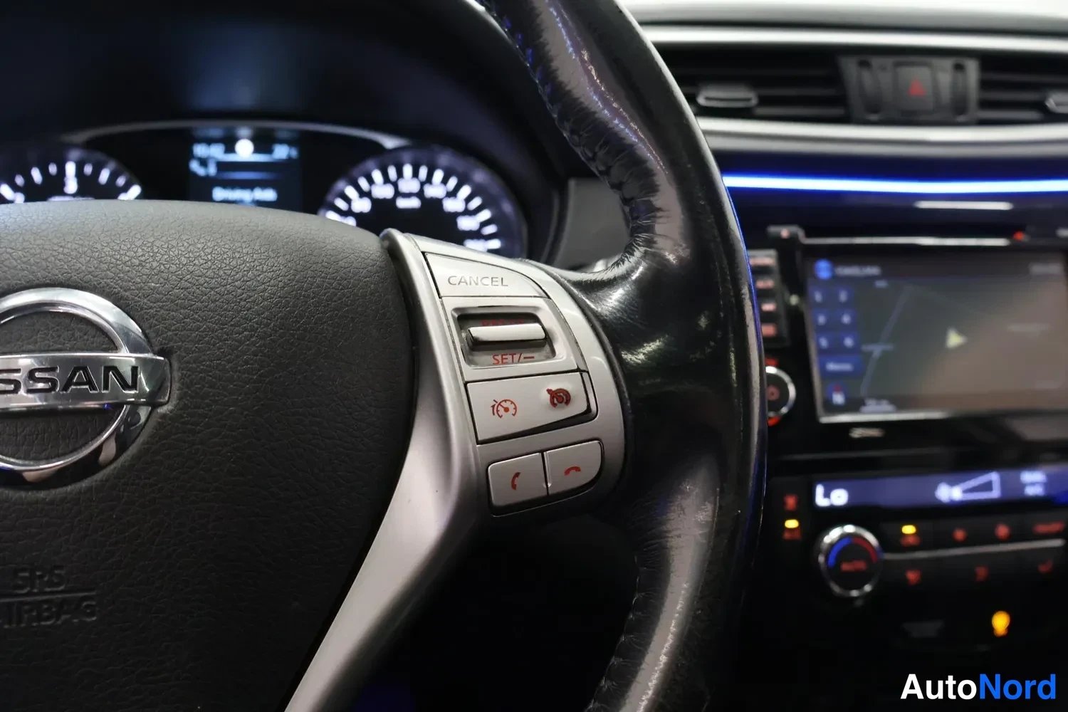 Sivulta: Sisäkuva ohjauspyörästä: Nissan-merkki, SRS AIRBAG -teksti, vakionopeudensäädön CANCEL- ja SET/-painikkeet sekä puhelinpainikkeet; taustalla mittaristo, sininen
