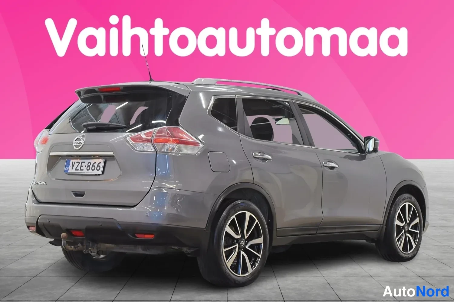Takaviisto: Harmaan Nissan X-Trailin takaosio ja oikea sivu kuvattuna kolmesta neljäsosasta; rekisterikilpi, kattokaiteet ja vetokoukku näkyvät.