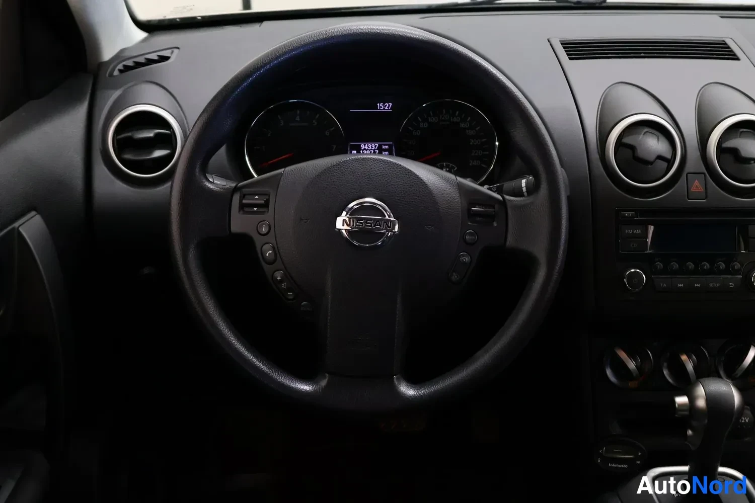Muu: Ohjaamon näkymä: Nissan-merkitty ohjauspyörä, kojelaudassa pyöreät ilmastointisuuttimet, radio ja automaattivaihteen valitsin.