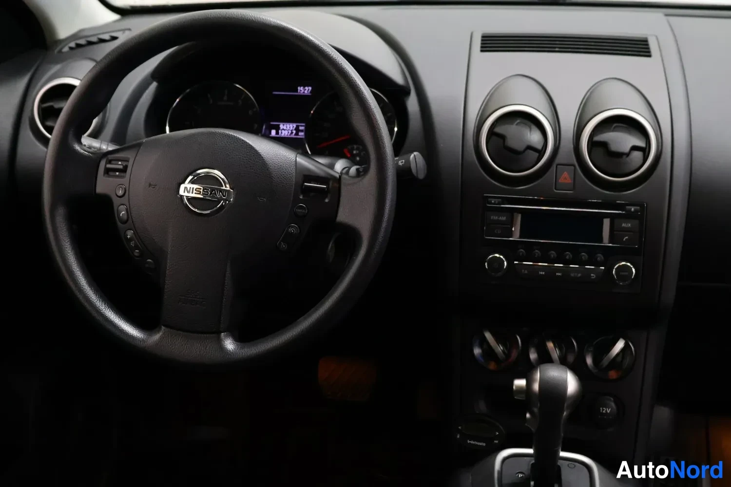 Muu: Ohjaamo edestä vasemmalta: Nissan-ohjauspyörä, mittaristo näyttää 94337 km, keskikonsolissa radio ja automaattivaihteen valitsin.