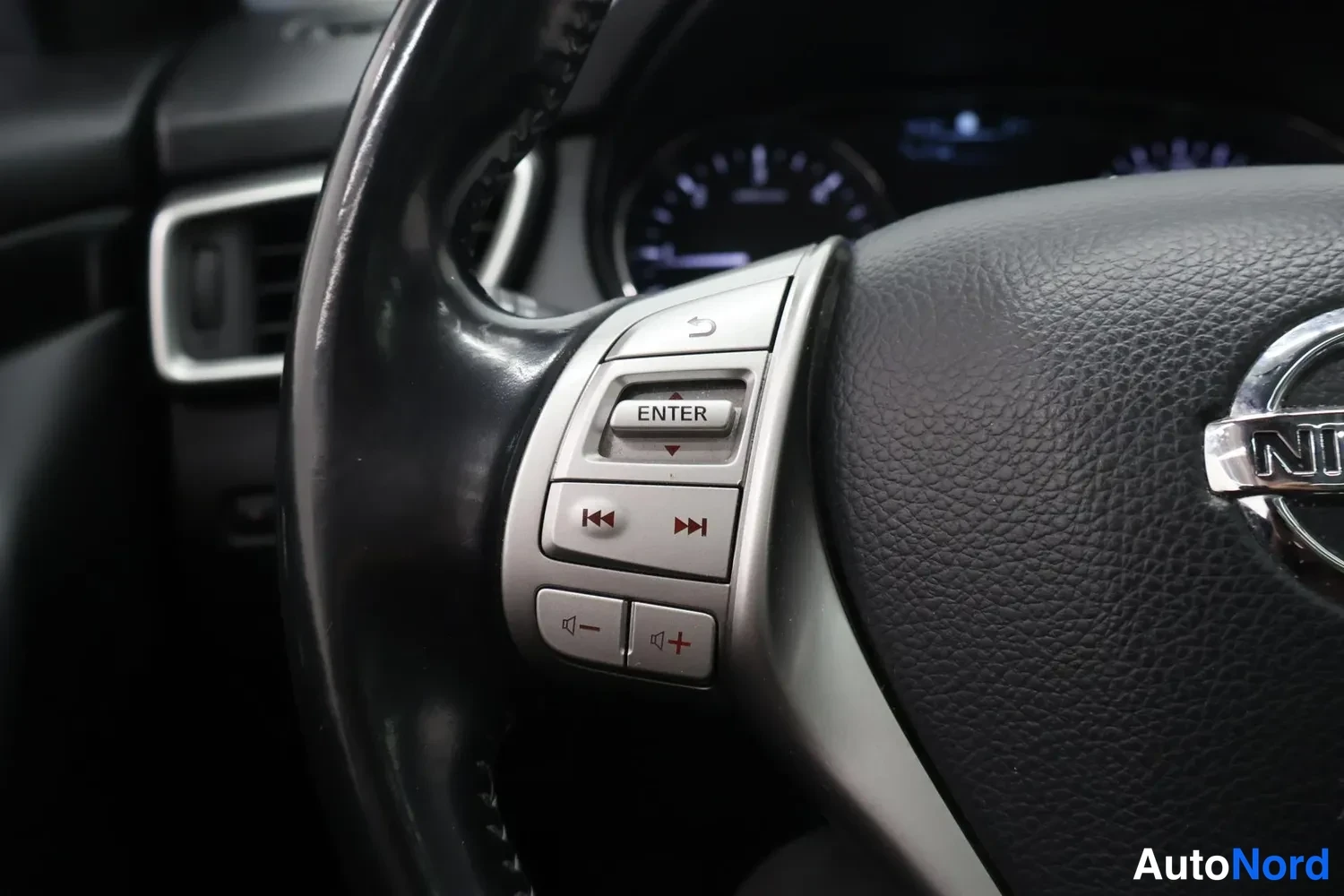 Muu: Lähikuva ohjauspyörän vasemmasta puhelin-/multimedia-osasta: ENTER-, äänenvoimakkuus- ja musiikinpainikkeet, Nissan-merkki näkyy osittain oikealla, kojelauta su