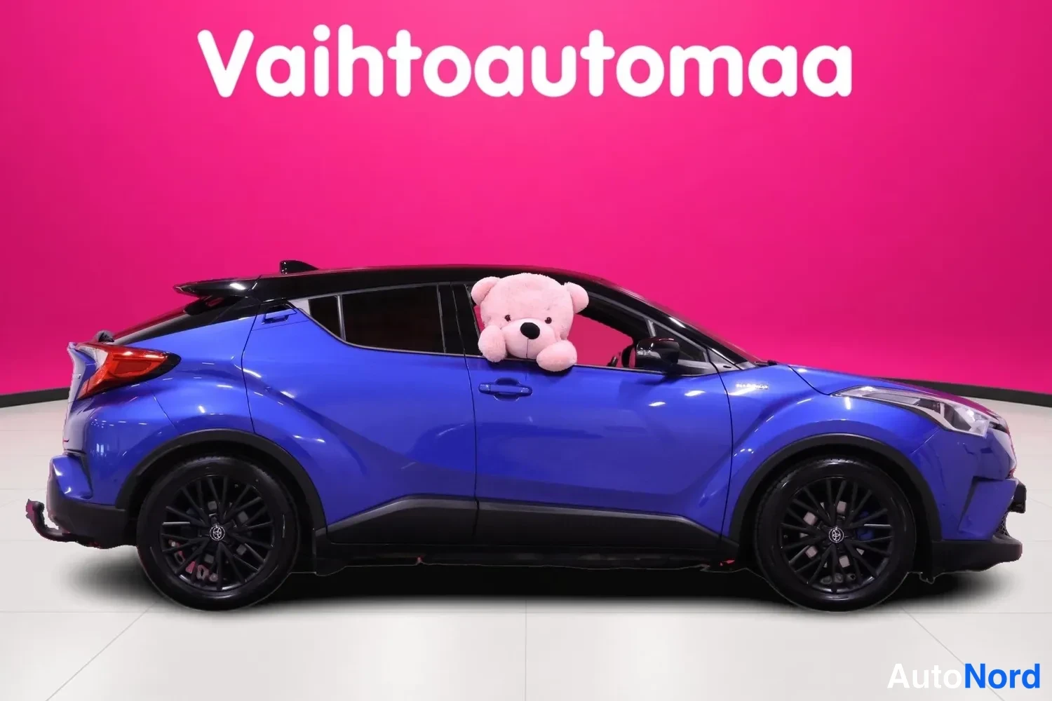 Etuviisto, nokka oikealle: Sininen Toyota C-HR etukulmasta, keula oikealla, mustat vanteet ja musta katto, vaaleanpunainen tausta.