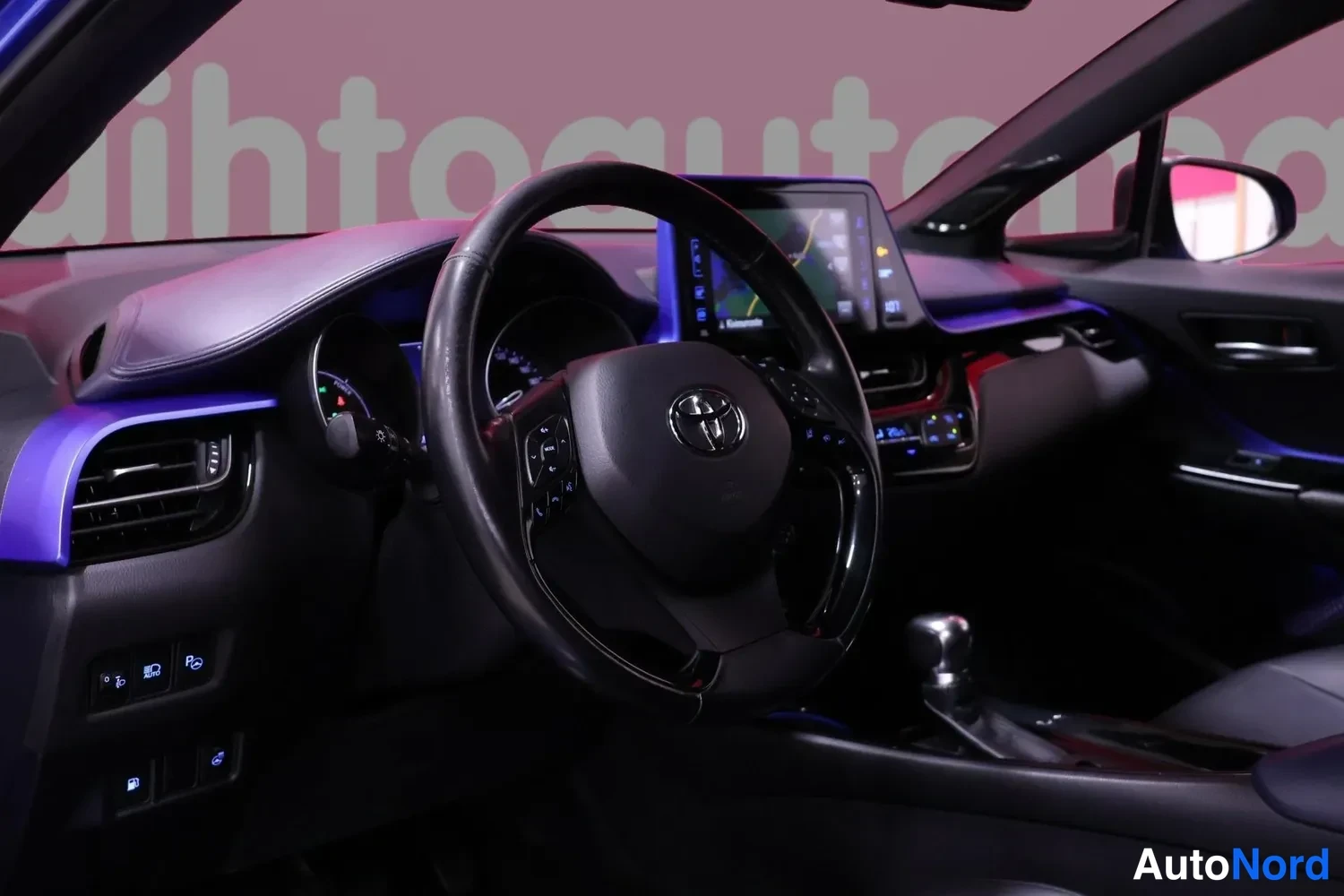 Yksityiskohta: Toyota C-HR:än etusohva: musta ohjauspyörä Toyota-logolla, digitaalinen mittaristo ja keskusnäyttö jossa kartta; tunnelmavalaistus sinisenä.
