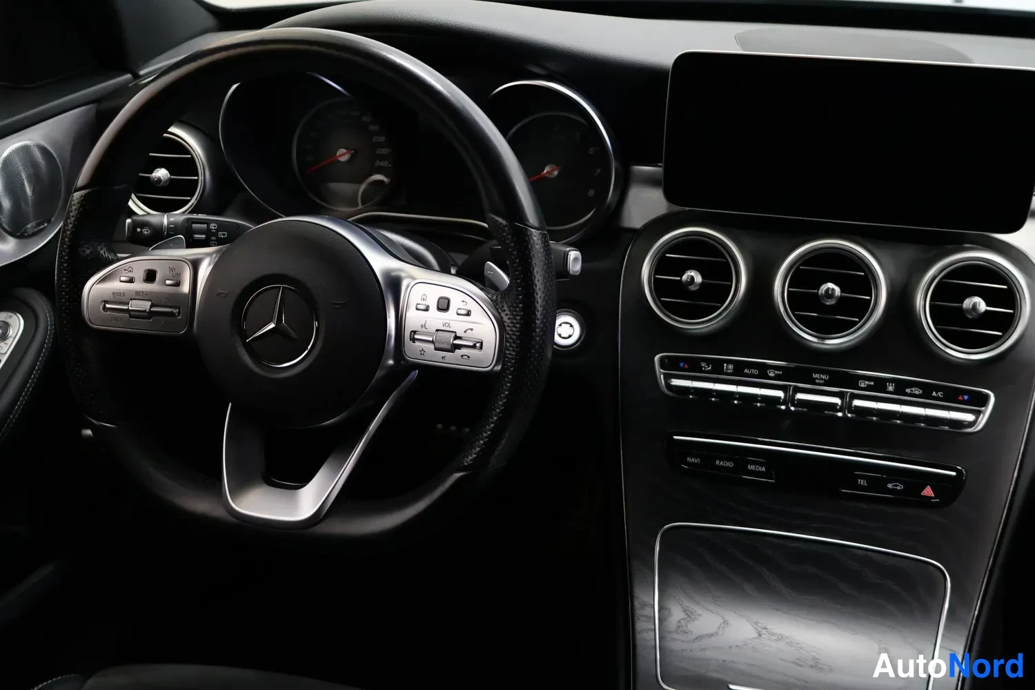 Muu: Kuljettajan puolen näkymä Mercedes-Benzin ohjauspyörästä, mittareista ja kosketusnäytöstä, jossa kolme pyöreää ilmastointisuutinta ja elektroniset painikkeet.