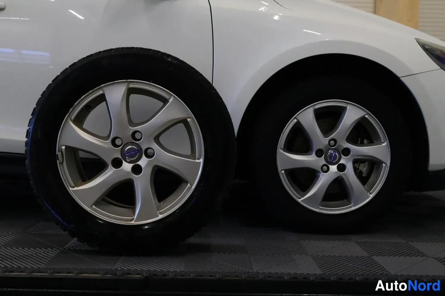 Sivulta: Valkoisen auton etupyörä ja vieressä nojallaan oleva varapyörä Volvo-merkkisillä vanteilla.