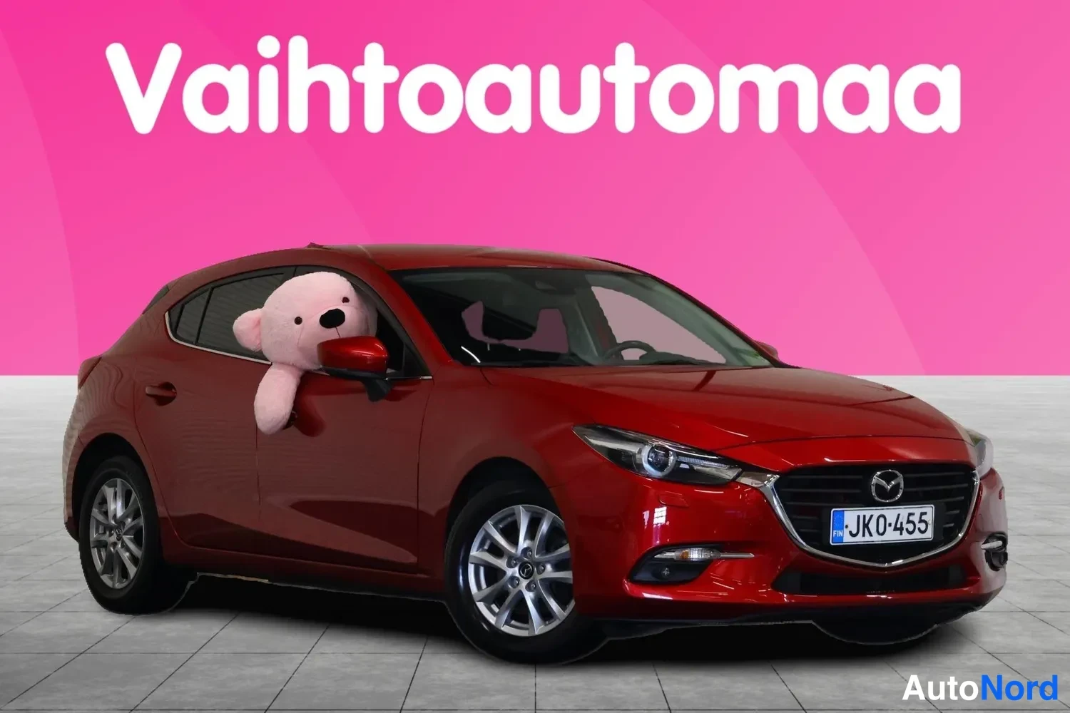 Etuviisto, nokka oikealle: Punainen Mazda 3 -henkilöauto edestä vasemmalta kuvattuna, rekisterikilpi JK0-455, pinkki tausta.