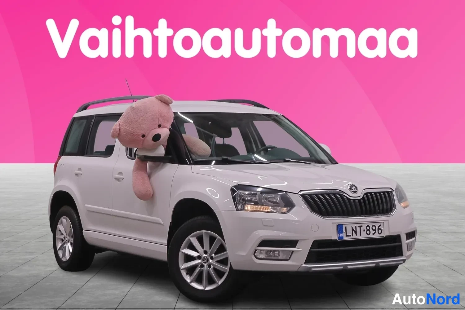 Etuviisto, nokka oikealle: Valkoinen henkilöauto kolmannelta etukulmalta, rekisteritunnus, etupuskuri ja kylki näkyvissä pinkkiä taustaa vasten.