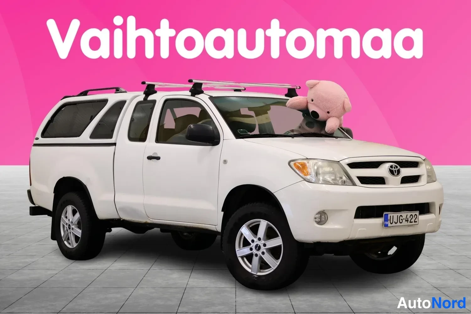Etuviisto, nokka oikealle: Toyota Hilux Extra 2006 -funga etuviistossa oikealta valkoisena, kattotelineet ja lainen kuormatila kannella näkyvissä.