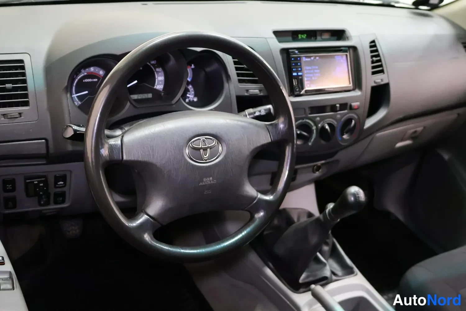 Edestä: Ohjaamon etukuva: Toyota-ohjauspyörä ja manuaalivaihteisto; kojelaudan keskellä näytöllä lukee 9:42.