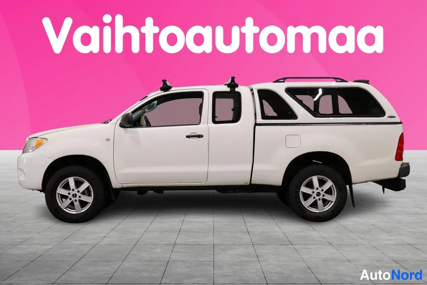 Sivulta: Valkoinen Toyota Hilux avolava sivulta vasemmalta, katolla telineet ja lavaosassa korokekate.