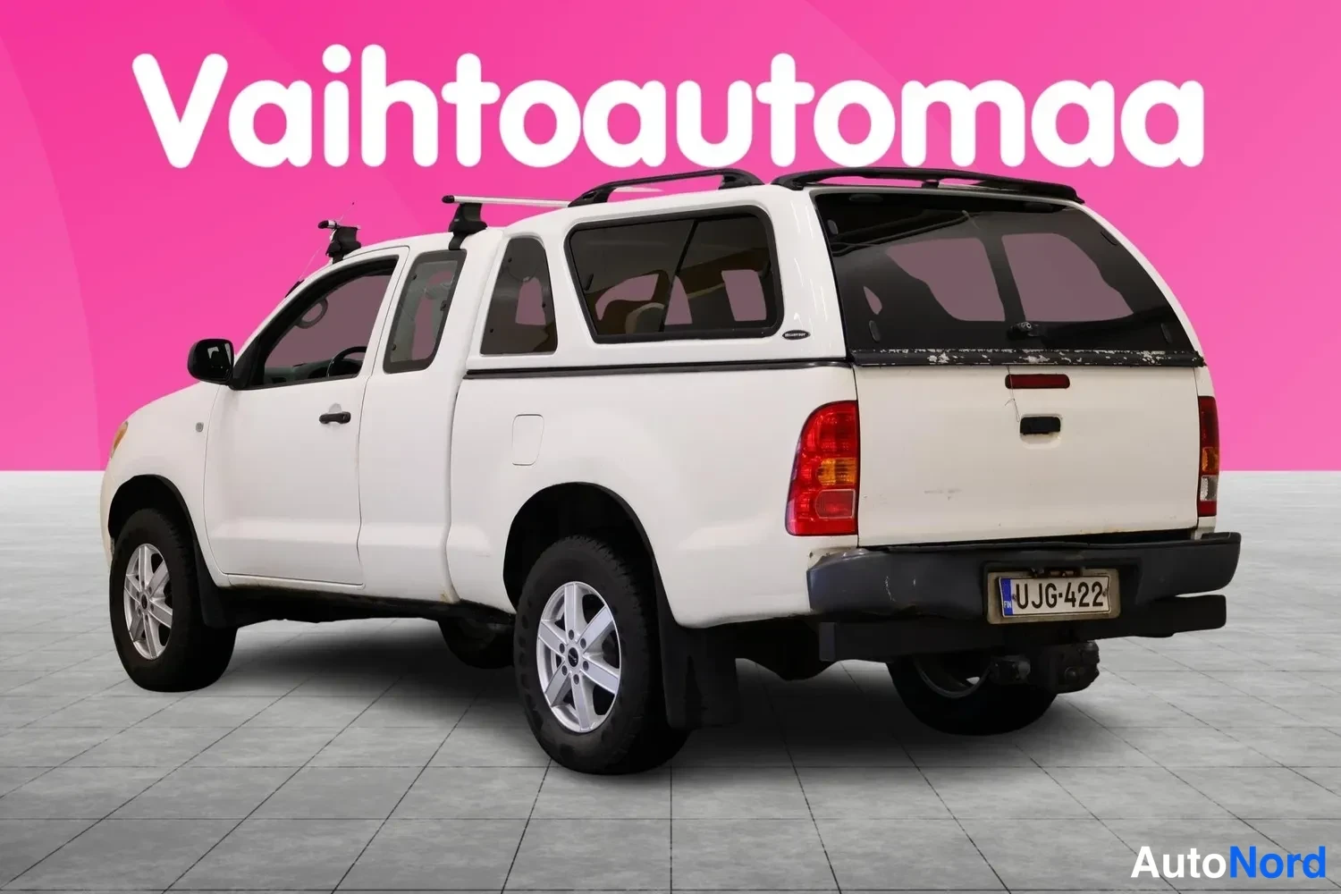 Takaviisto: Valkoinen Toyota Hilux avolava takaviistosta vasemmalta, lavaosassa korokekate ja katolla telineet.