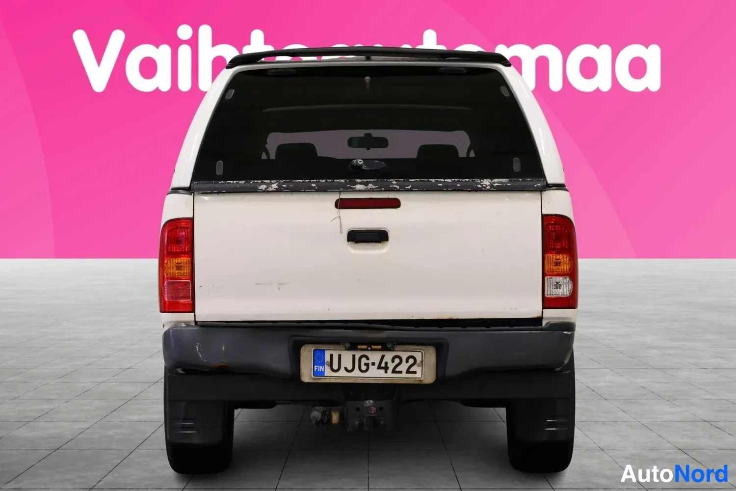 Takaa: Valkoinen Toyota Hilux avolava suoraan takaa päin, rekisteritunnus ja korokkeen takalasi musta.