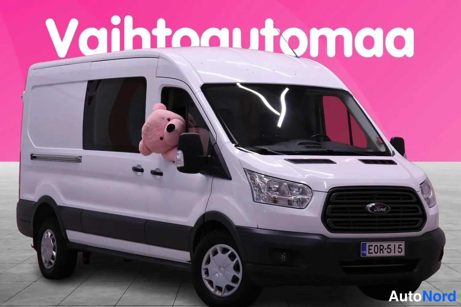 Etuviisto, nokka oikealle: Valkoinen Ford Transit -pakettiauto etuviistöstä oikealta, rekisterikilpi, vaaleansävyinen pehmolelu etuikkunassa.