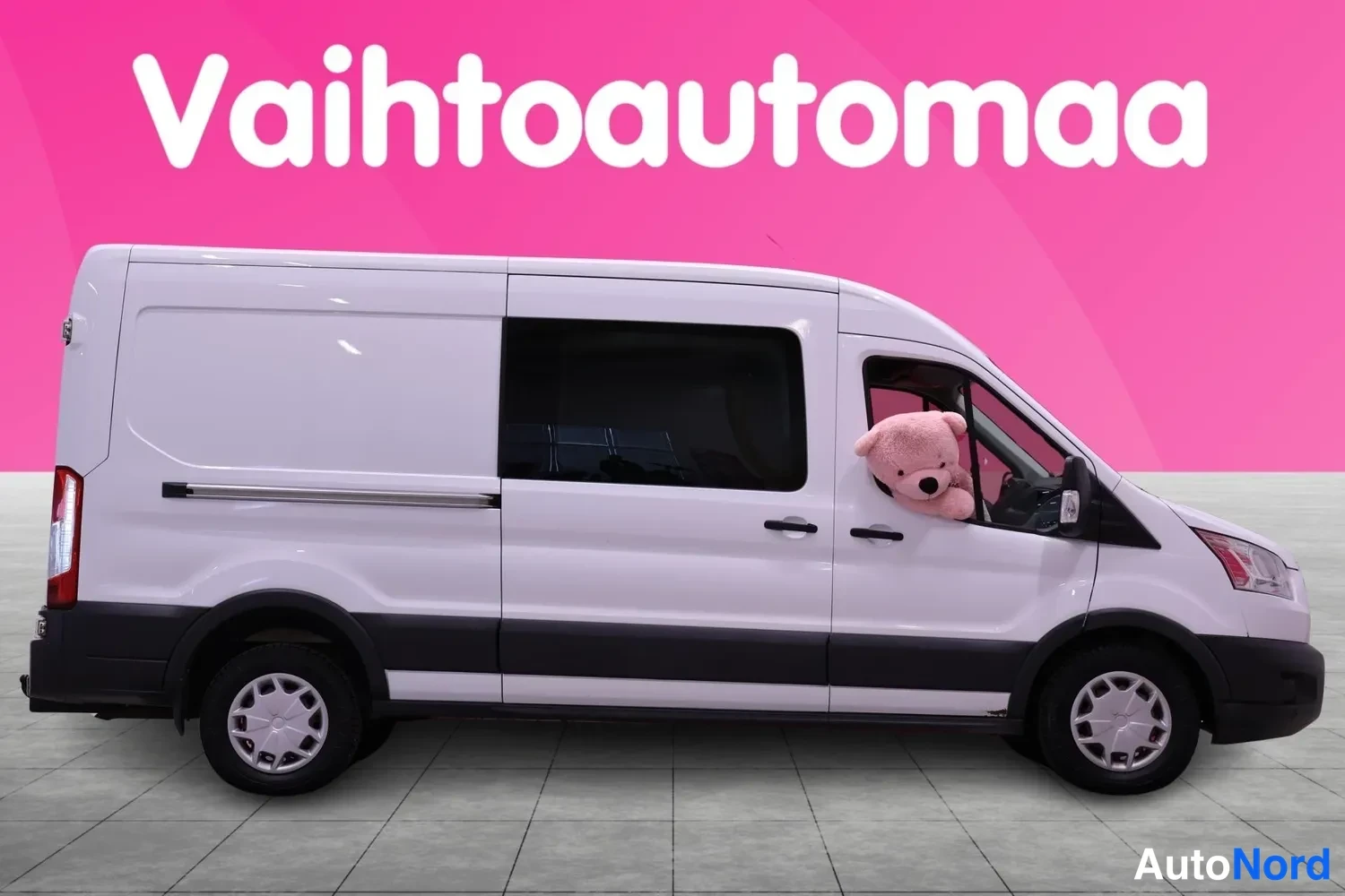 Etuviisto, nokka oikealle: Valkoinen Ford Transit -pakettiauto sivulta oikean etukulman näkyessä, tummat takaoven ikkunat ja pehmolelu kuljettajan ikkunassa.