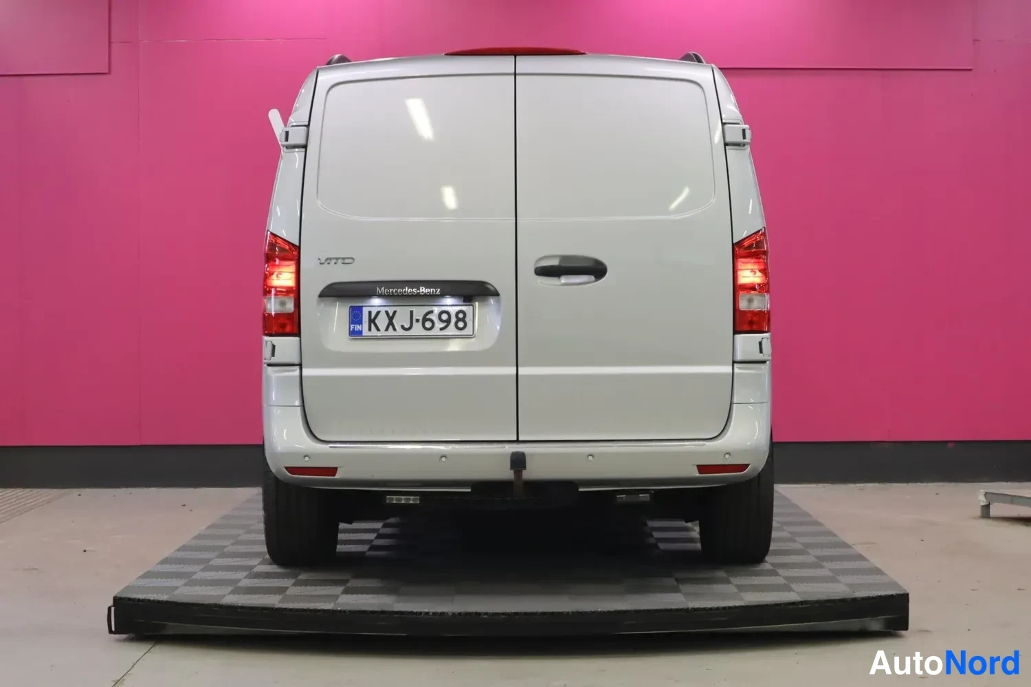 Takaa: Mercedes-Benz Vito -pakettiauton takaosan suora näkymä: kaksiosaiset takaportit, rekisterikilpi ja harmaa metalliväri vaaleanpunaisen taustan edessä.
